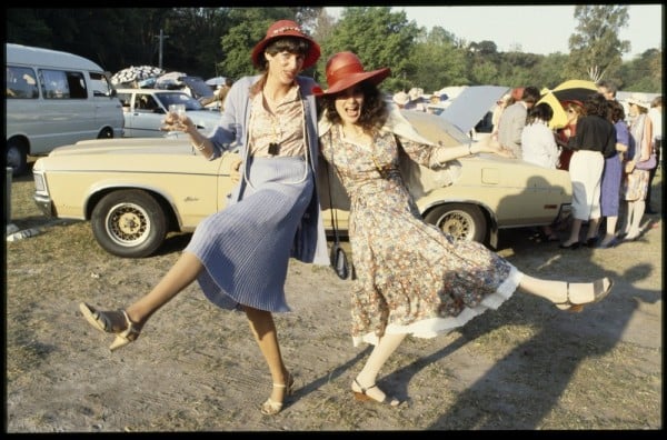 Two women, both wearing red straw hats, woman on left holding a drinking glass, cars parked behind them, most with boots open, beach umbrellas and small gatherings of people.