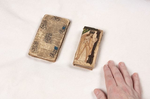Two small books from the John Emmerson Collection with embroidered bindings on a white pillow. A librarian's right hand is next to the item on the right for scale. The items are smaller than the size of a palm. 