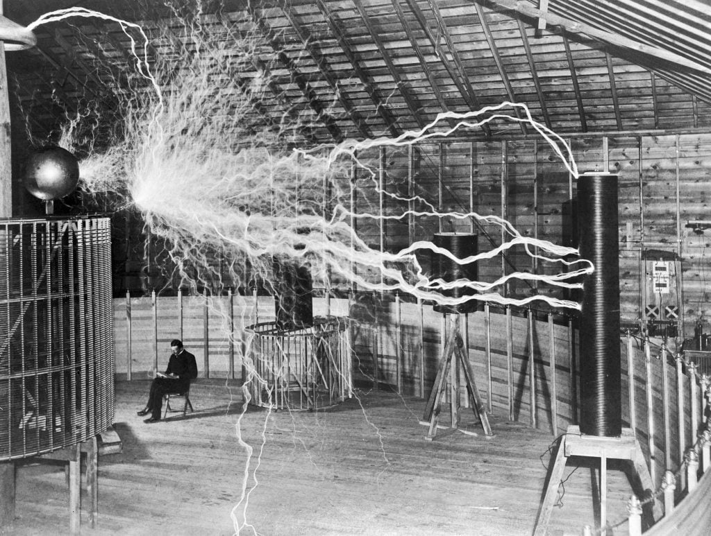 Nikola Tesla sitting with his equipment.