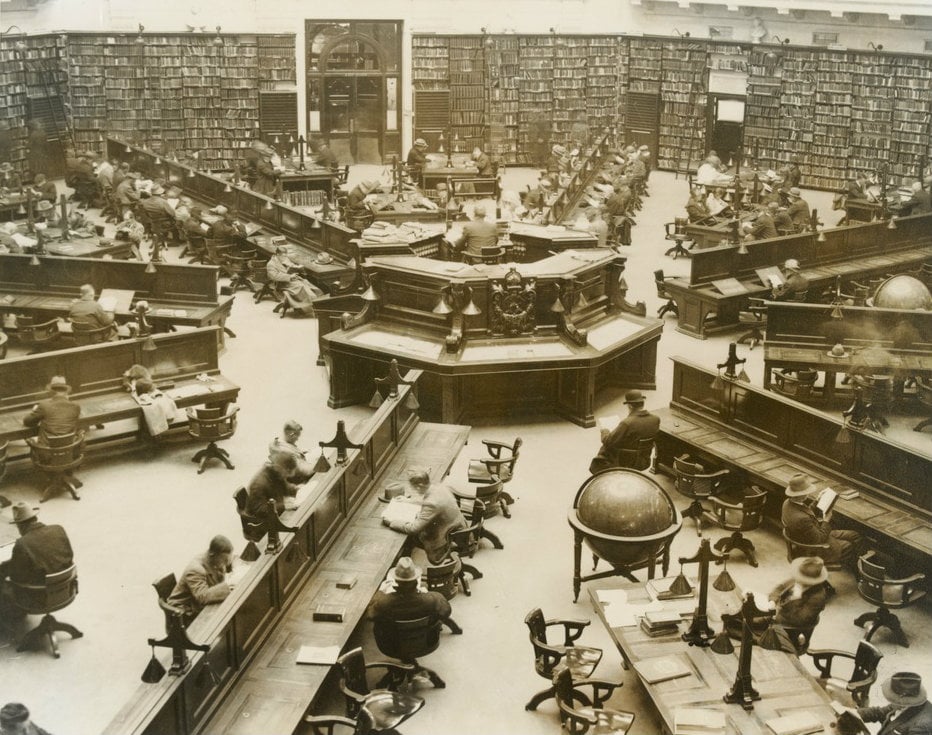 Photograph of the Dais in the Domed reading room, showing library attendant in centre, with mirror above desk. Desks radiate out from the Dais like spokes. The shelves on the walls are stacked high with books.