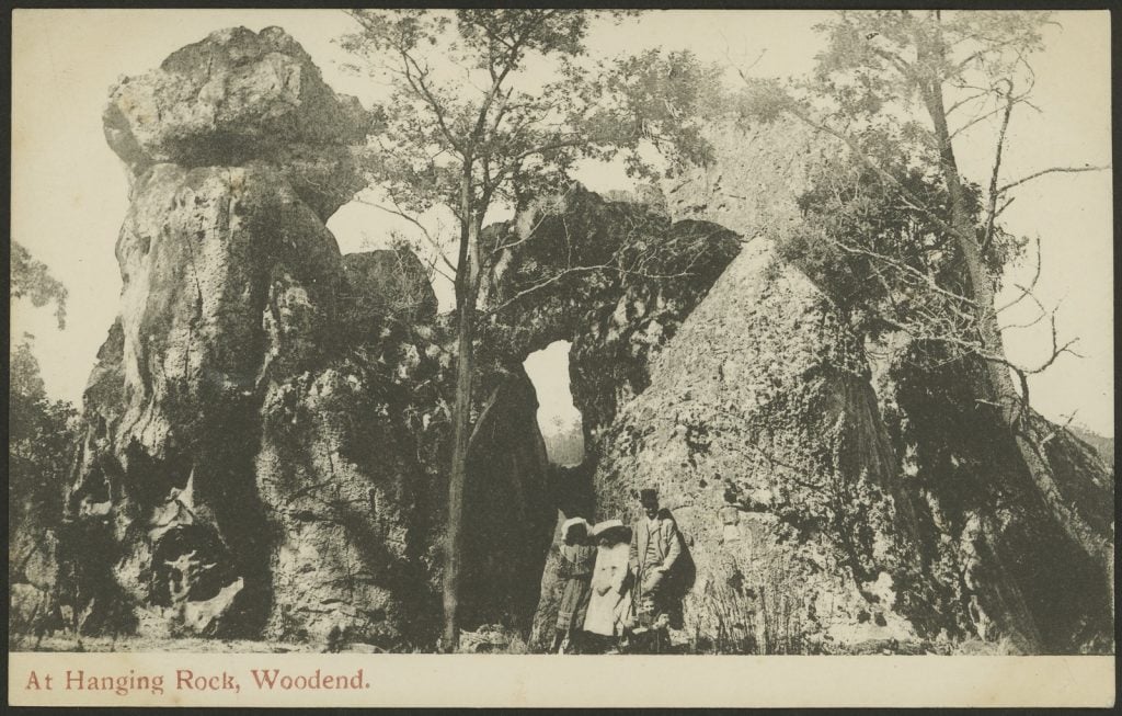 Black and white postcard depicting two ladies and a man wearing early twentieth century dress standing in front of Hanging Rock
