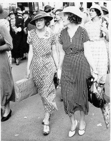 Black & white photograph of two women, whole-length, wearing hats and long dresses, carrying bags and parcels, walking through crowded street.