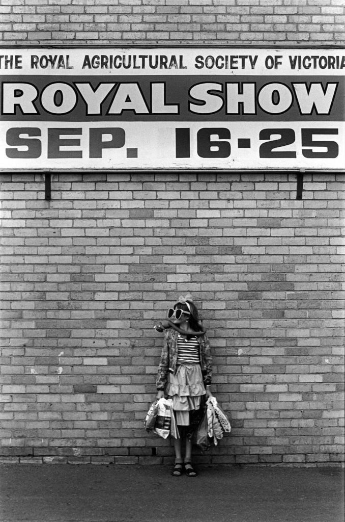 Black and white photo of the photographer's daughter, Sarah Maddison, standing beneath a sign advertising the Royal Melbourne Show, September 16-25 [1982]. Sarah wears outsize sunglasses with white frames, a long balloon worn as a scarf round her neck, and she carries many showbags.