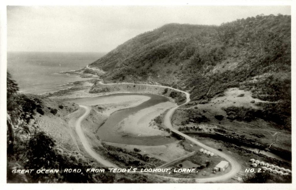Photograph, view from elevated point of campsite and winding road, a creek running under a bridge and out to sea.
