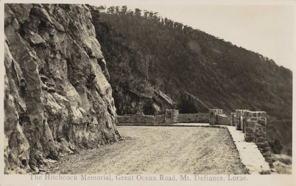 photograph, cut rock face along side the road, with a stone wall constructed on the sea side of the road. wooded hills beyond