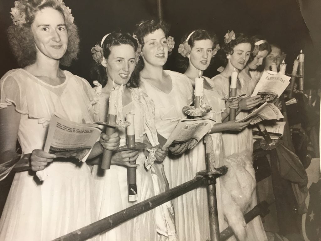 Seven women in white dresses, holding candles