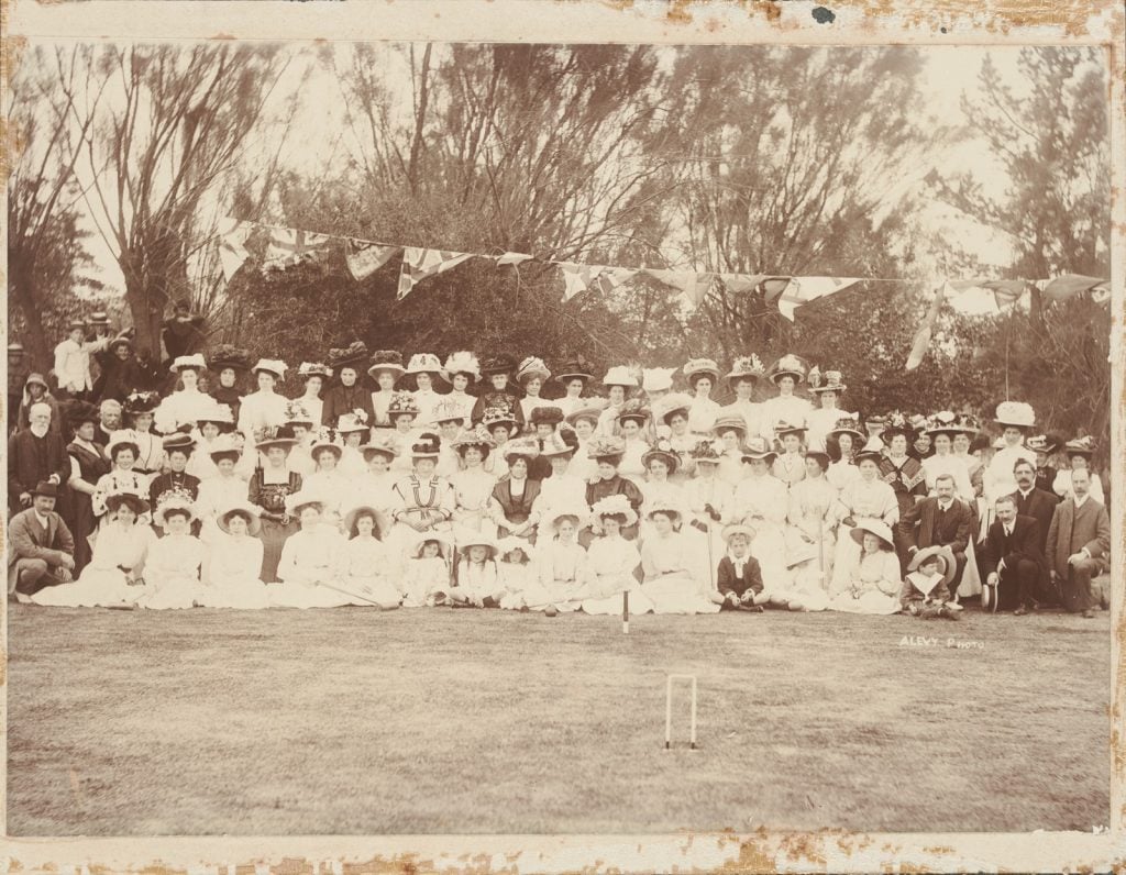 photograph of a large group of mostly women, and some men., The women dressed mostly in white, with large hats. a string flying flags suns across the middle of the photo, a croquet hoop in the foreground