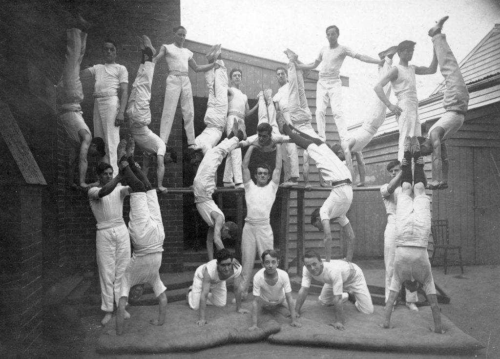 men and boys creating a human mountain, perched and supporting each other in formation. wearing white trousers and singlets or vests.