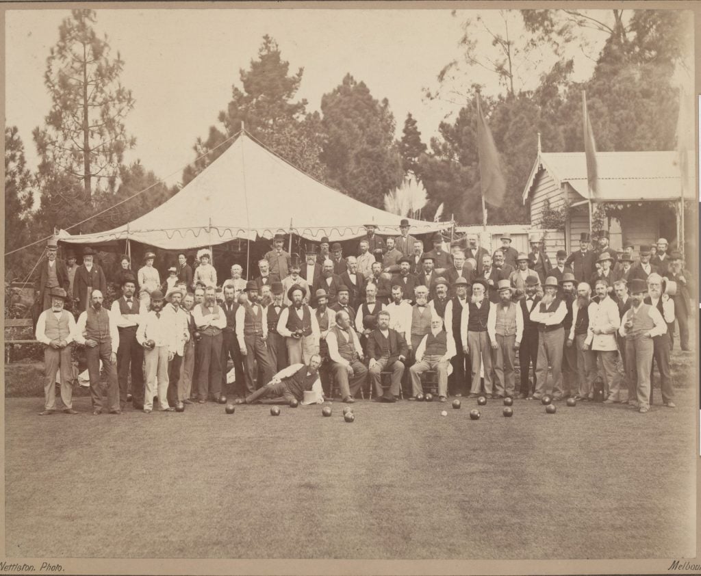 Picture of a large group of men bowlers standing,a few seated and one reclining in front of a marquee tent,  with a building to the right, with trees beyond, and a clump of pampas grass. There are some bowling balls scattered on the green in front of them 