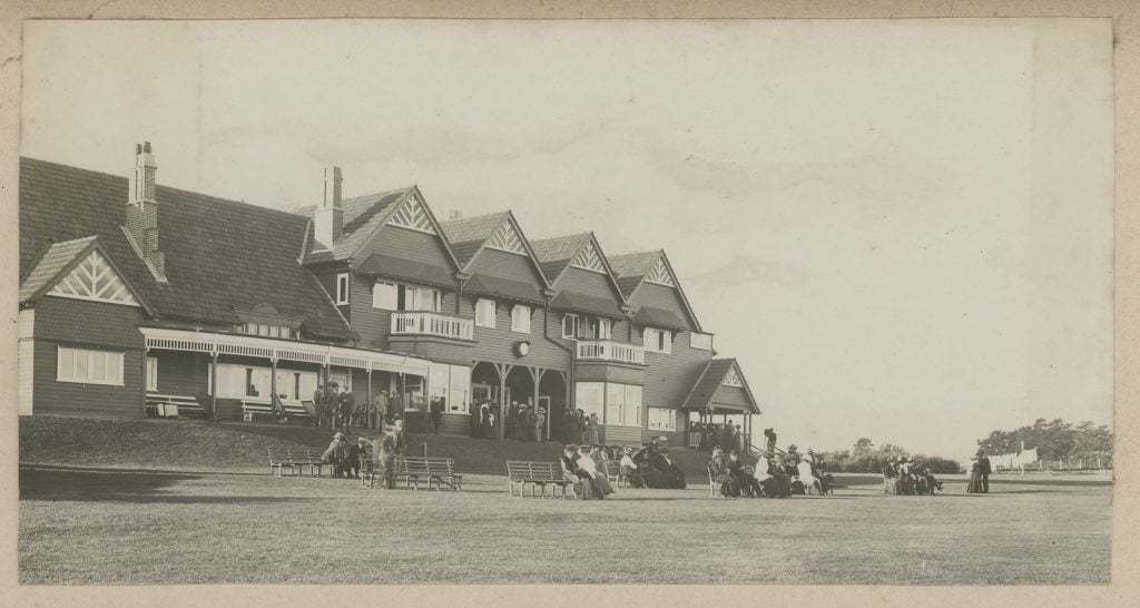 Photograph of the club rooms a grand double story building with gables rooves, people sitting on timber benches in the foreground