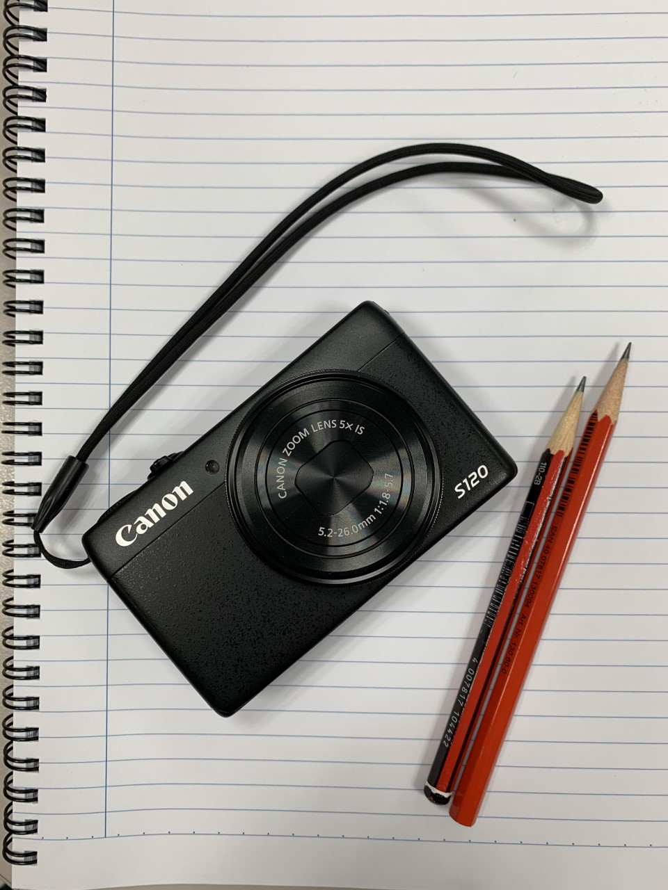 Digital camera sitting on top of notebook with pencil