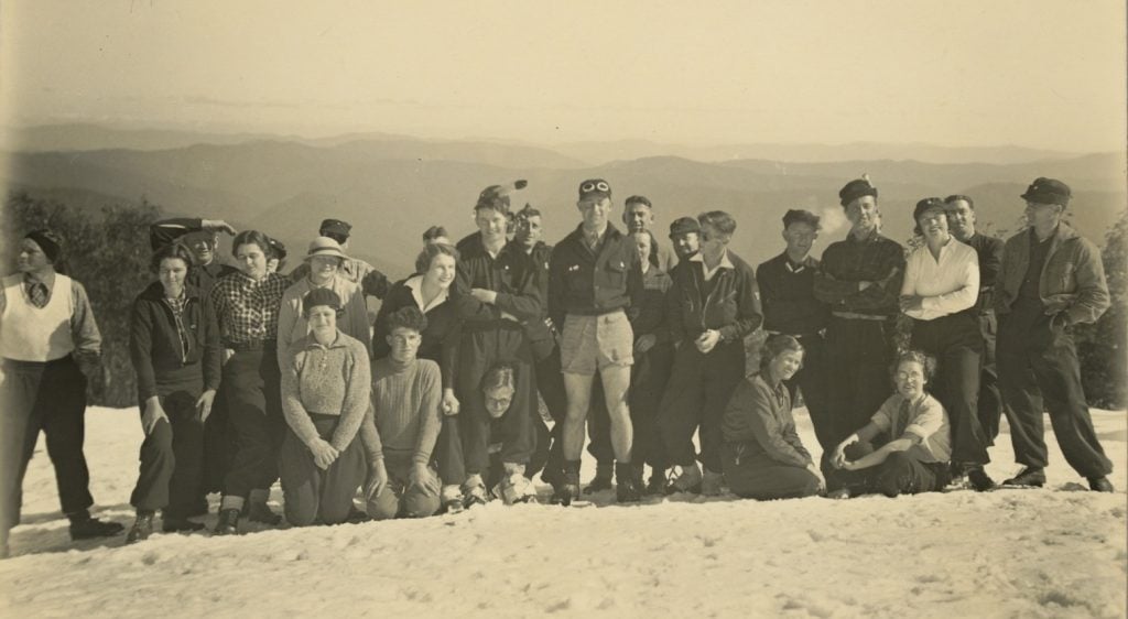 group photograph of skiers standing and kneeling in the snow, a sunny date, with receding mountains in the background. Some wearing shirtsleeves, and one man in shorts