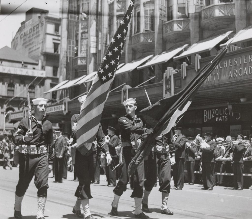 American sailors marching past crowds lining streets of Melbourne: U.S. Navy standard bearers with Stars & Stripes and Navy flag; U.S Marines in armored vehicle passing Town Hall in Swanston Street; Crowd lining street as U.S. servicemen march past.