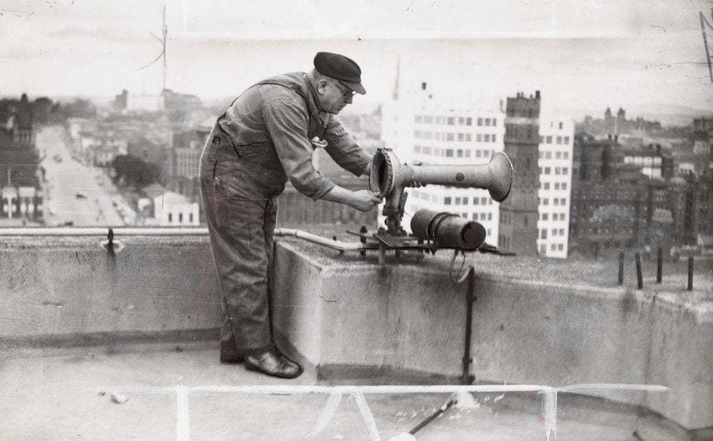 Shows man wearing overalls, with a peaked cap and rolled up shirtsleeves, bending over an air raid siren, which is on the roof of a tall city building