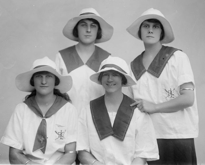 Four young women, full face, two seated with two standing behind them. Subjects, from Bendigo Ladies' Rowing Club are: J. Hesse, N. Oldfield, T. Walker, J Weibgen. All wearing white short sleeved blouses, three with sailor style collars, one with kerchief, and wide brimmed hats. Two women have an insignia on the breast pocket of their blouses PLC with crossed oars.