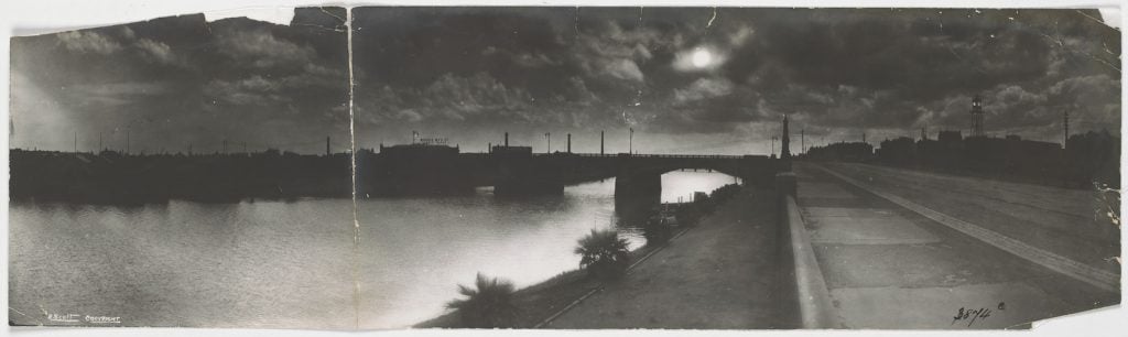 panoramic photograph taken on moonlight looking along a river, with palm trees along a bank, a promenade and looking towards a bridge, with the moon shining through clouds.