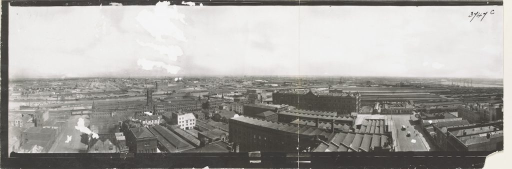 Panoramic photograph overlooking factories with railway lines and harbour with ships masts in the background.