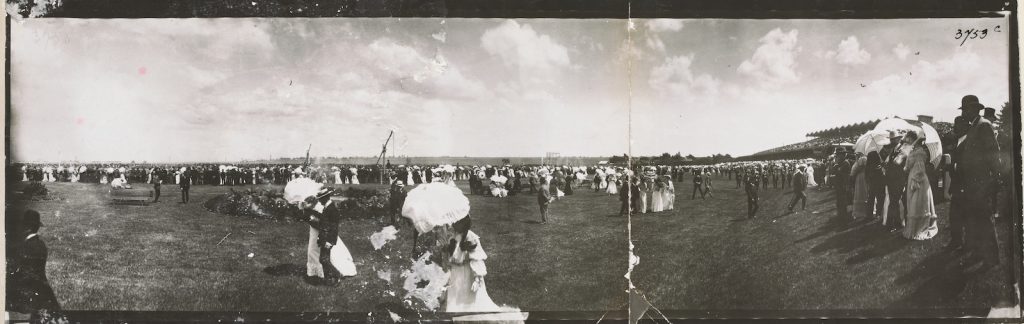 Panoramic photograph taken on a day the races, women dressed up, many carrying parasols and men in suits and hats promenading on the lawns with the grandstand to the rear right side.