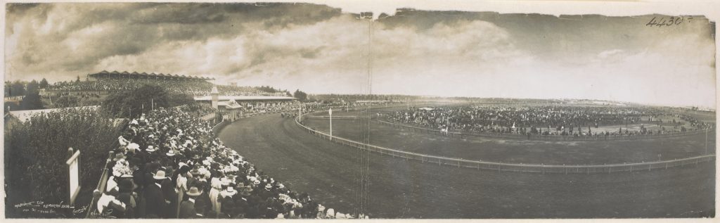 panoramic photograph taken at Flemington racecourse with crowds watching the race, grandstand to the left and the track curving around in the foreground of the photograph.