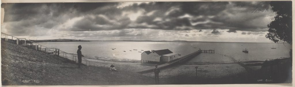 panoramic photograph, a moonlit night three people in the foreground with boat sheds, a jetty and the lake beyond.