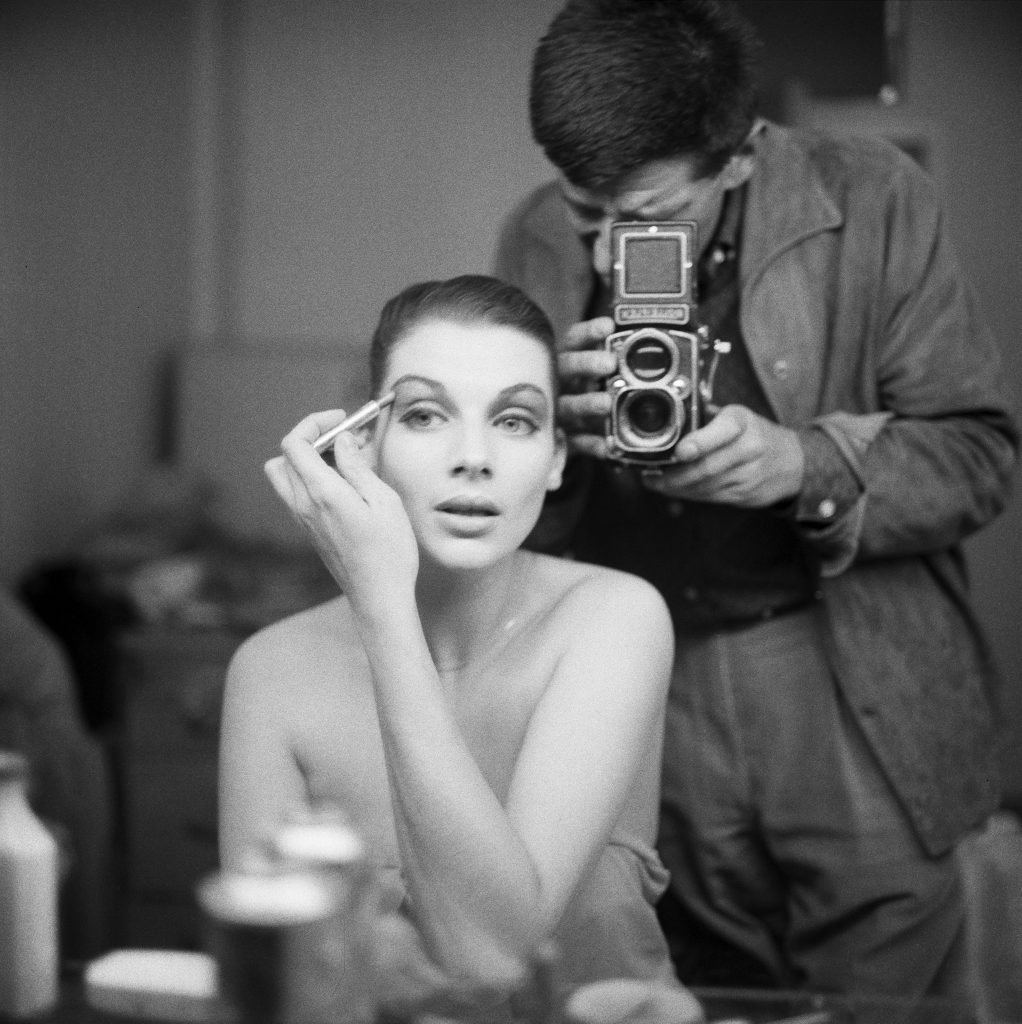 Photograph of model Maggie Tabberer seated in front of a mirror putting on makeup, photographer visible in mirror reflection, standing behind her, taking her picture, likely Henry Talbot.