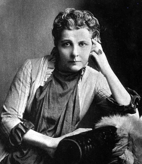 Photograph of Annie Besant, born 1849, died 1933.. She sits with a look of determination on her face, wearing a dress with frilled sleeves and a scarf.  Her hair is pulled back, with thick curls on her head.  