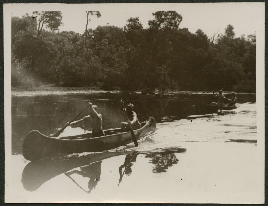 Two teenage boys rowing a canoe at the beach, churning the foam. Another canoe follows. It is very sunny. The canoe and rowers are reflected in the water. There are trees in the background. 