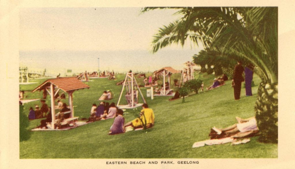 Lawns at Eastern Beach showing people sitting on the grass, shelters, with a children's playground in the background. 