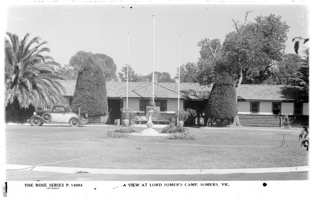 Long building with tiled roof and large trees behind. Large palm tree,  two large trimmed  trees and three flag poles in front. 1930s style car parked in front of  palm tree. Large lawn in foreground, with plants in centre and sprinkler. 