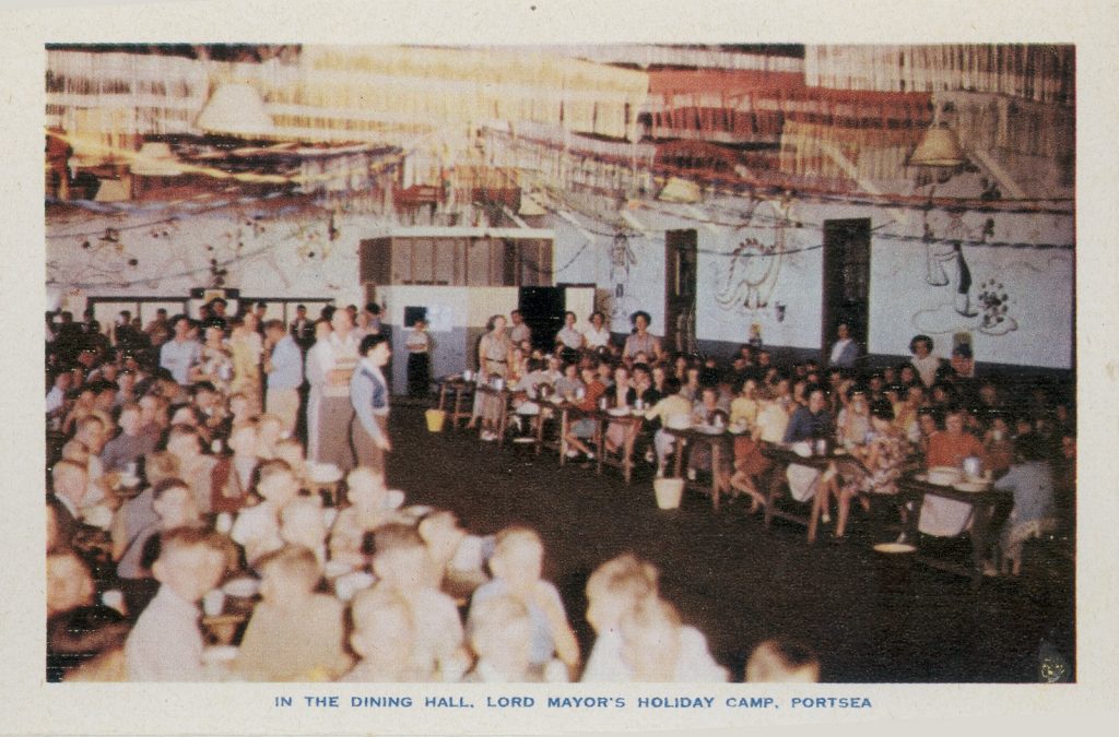Colour photo of large number of children and adults sitting at tables in the dining hall, Portsea camp. Some adults standing.  Large illustration of a dinosaur and other images on the wall.