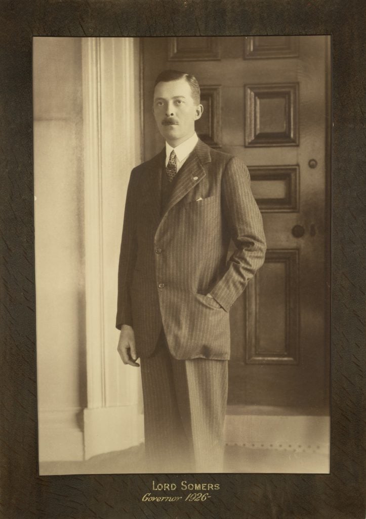 Lord Somers with moustache, wearing pin striped suit and patterned tie, standing with left hand in pocket.