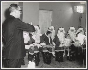 Black and white photo of a man in a suit addressing a room of Santas