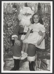Black and white photo a girl in a white dress sitting on Santa Claus' knee in front of a glittering backdrop