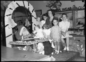 Black and white photograph of Santa sitting at a desk in a store, under an archway, with young children and a woman gathered around
