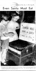 Black and white photo of a young boy and girl looking at a sign on Santa's chair that says "Santa has gone to lunch. Back at 1pm"