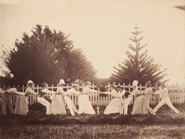 Sepia tinted photo of a women and men in late 18th century dress playing tug of war in a garden