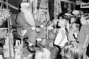 Black and white photo of Santa in a store, sitting on a chair upon a dais, surrounded by young children
