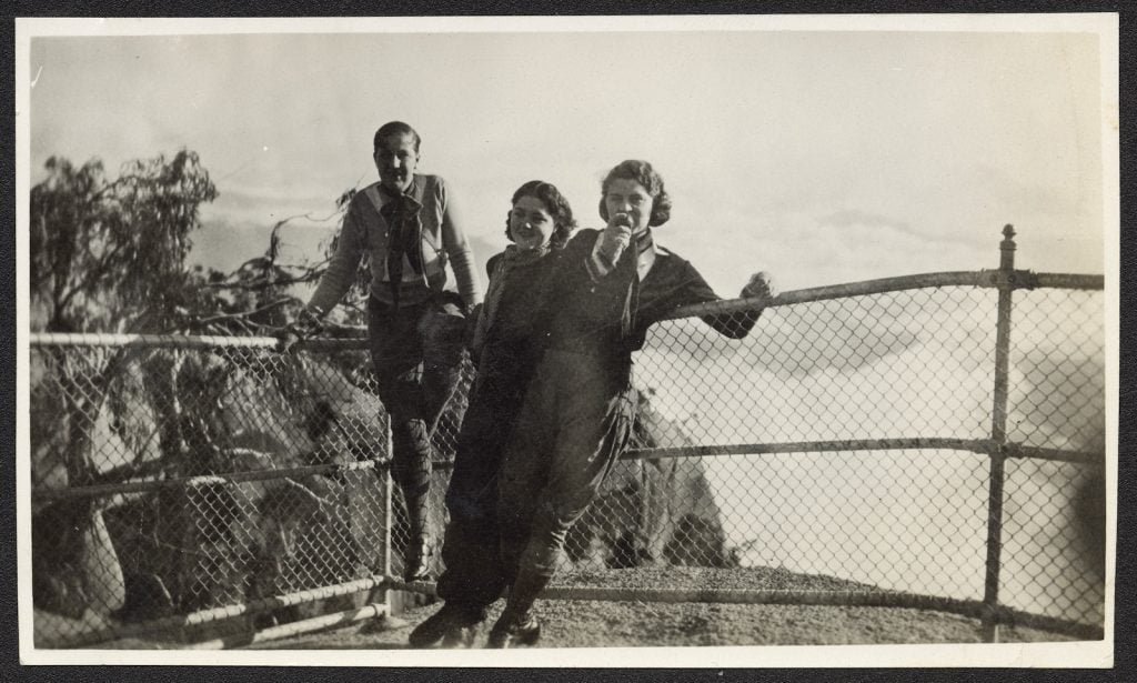 Three young people, a man and two women, stand in front of a wire fence at the lookout, Mount Buffalo. They wear walking clothing.