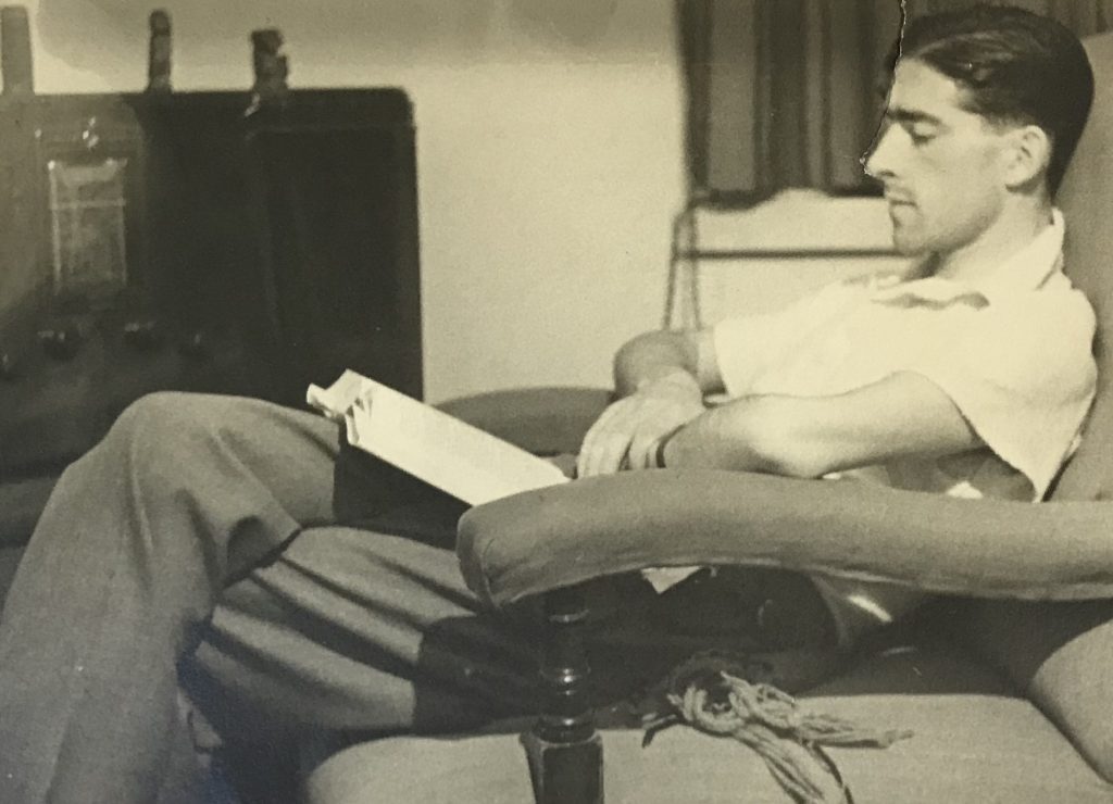 Black and white photo of Laurie Thomas  reclnging in a chair with an open book on his knee 