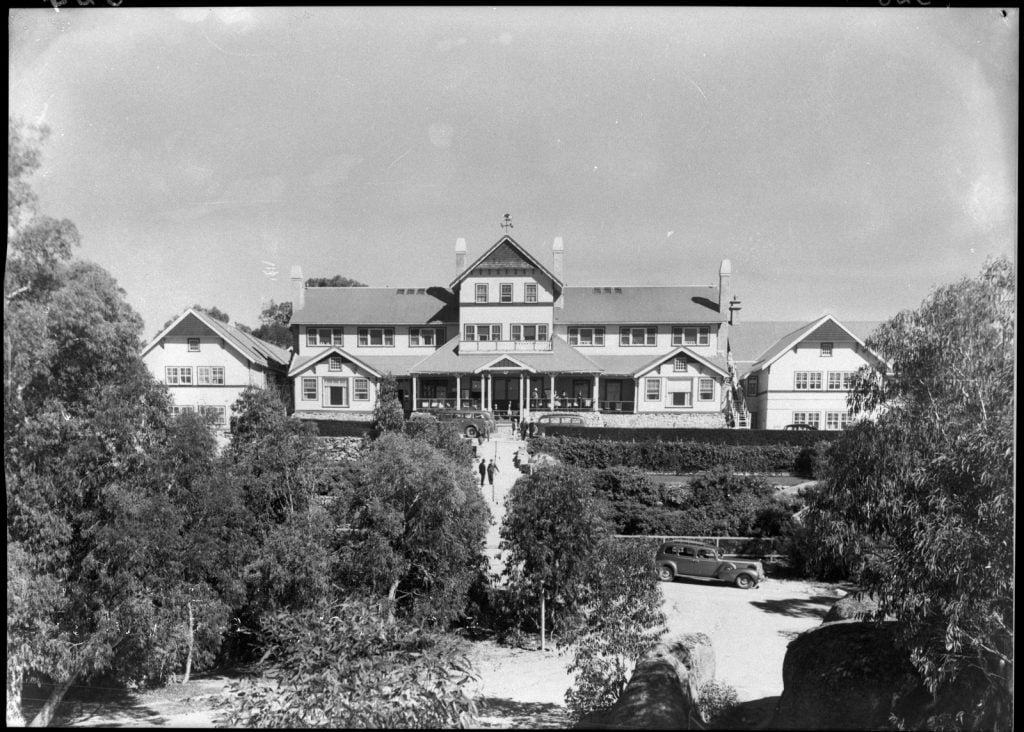 Photo of Mount Buffalo Chalet, a large building with gables and a verandah. People walk up the long path  to the verandah. Eucalypt trees in the foreground.  