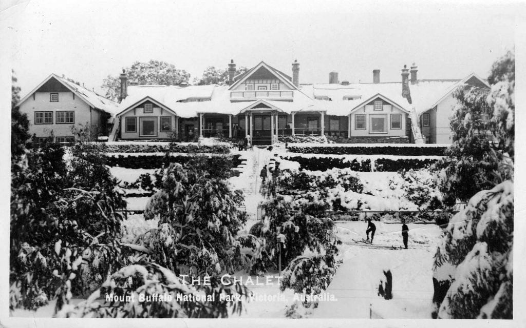 Mt Buffalo Chalet in the snow. The roof, path, ground and trees are covered in snow. Several people stand in the foreground. 