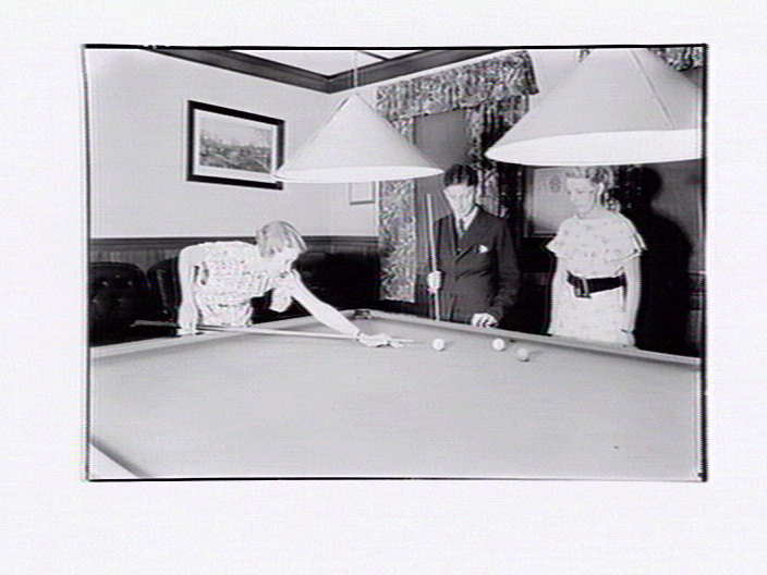Three people playing billiards. A woman aims a billiard  cue stick at a ball on the pool table while a man watches, holding a cue stick. Another woman watches.  