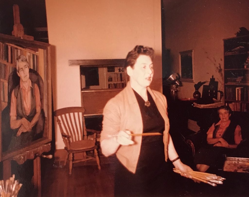 Colour photo of Lina Bryans holding paintbrushes with Nettie Palmer seated in the background. Lina's portrait of Nettie is on an easel to their left