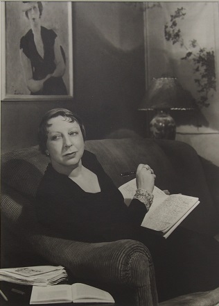 Black and white photo of Jean Campbell sitting on couch writing in a notebook, her portrait on the wall in the background 