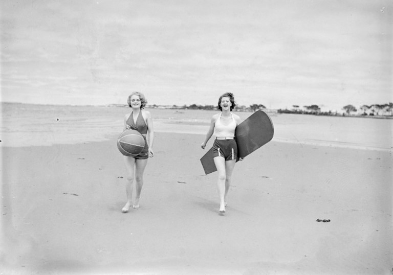 Two women in swimsuits on a beach, one carrying a beach ball and the other carrying a surf board
