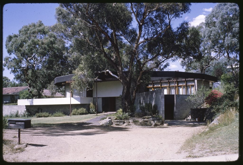 photograph, curved house building, brick walls flanking the main structure - view from the frontage, with large gumtree in the foreground, and letterbox in view.