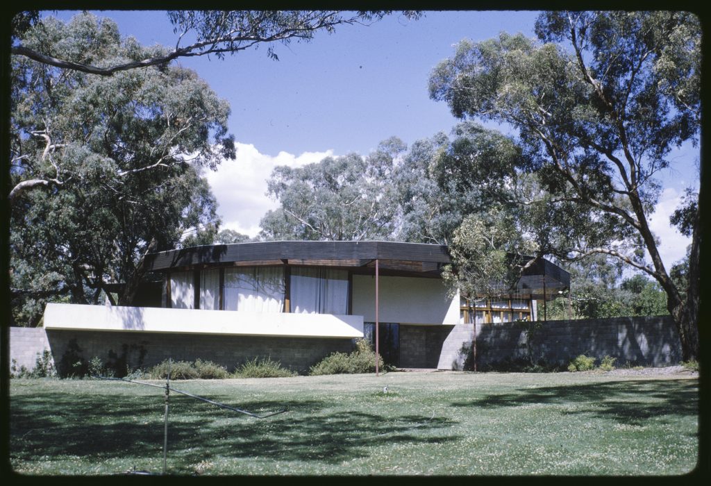 photograph of house - curved main structure flanked by brick walls, surrounded by gum trees.
