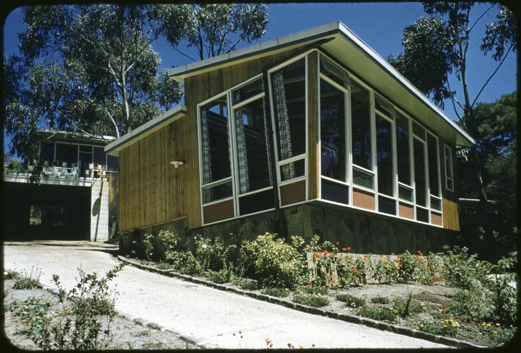 photograph of front section of a timber house with large windows, angled roof section,rear portion of house partly visible. Taken on a sunny day with gum trees behind the house. 