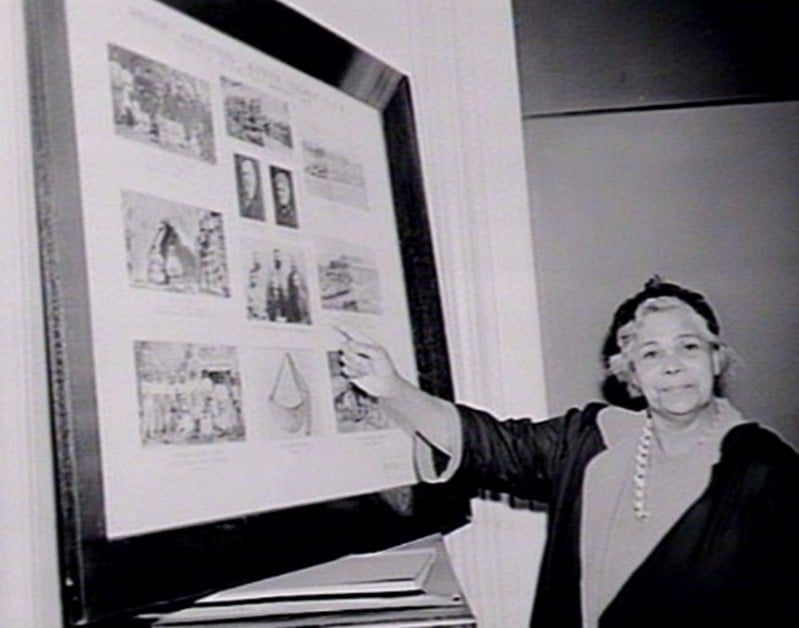 A photograph of an Aboriginal woman wearing a black gown and hat and pointing to a framed collection of pictures