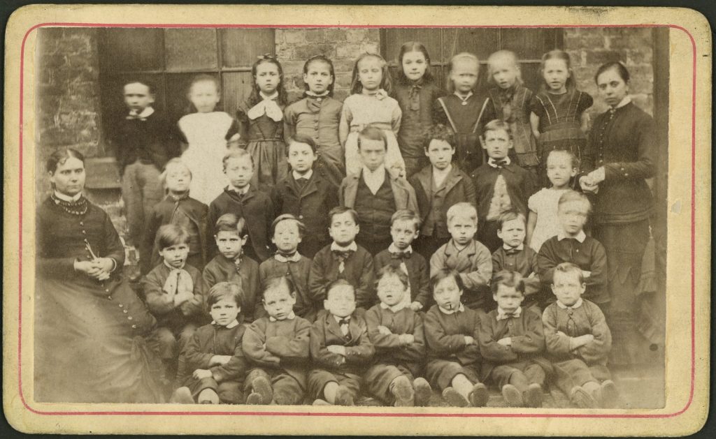 Shows four rows of children, girls standing at the back with a female teacher at each end.
